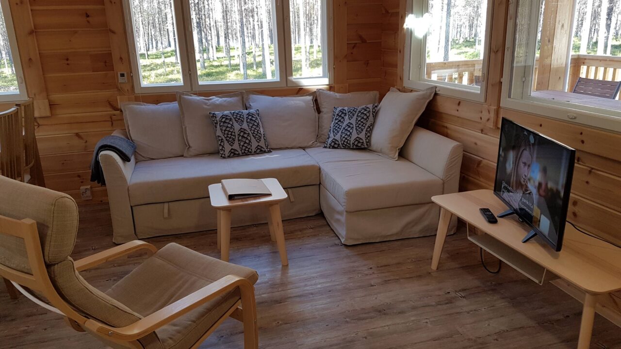 Lumon hirsimökin olohuoneessa vaaleanruskea divaanisohva ja nojatuoli, pieni sohvapöytä sekä televiso, ikkunoista näkyy mäntymetsään ja terassille.