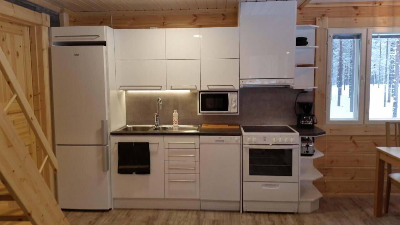 Lumon hirsimökin keittiö, jossa on valkoiset kaapistot, ja näkyvillä jääkaappi, liesi, mikroaaltouuni, kahvinkeitin ja leivänpaahdin.