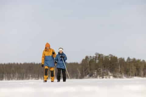 Kaksi henkilöä hiihtää perinteisellä hiihtotyylillä järven jäällä, horisontissa metsä.