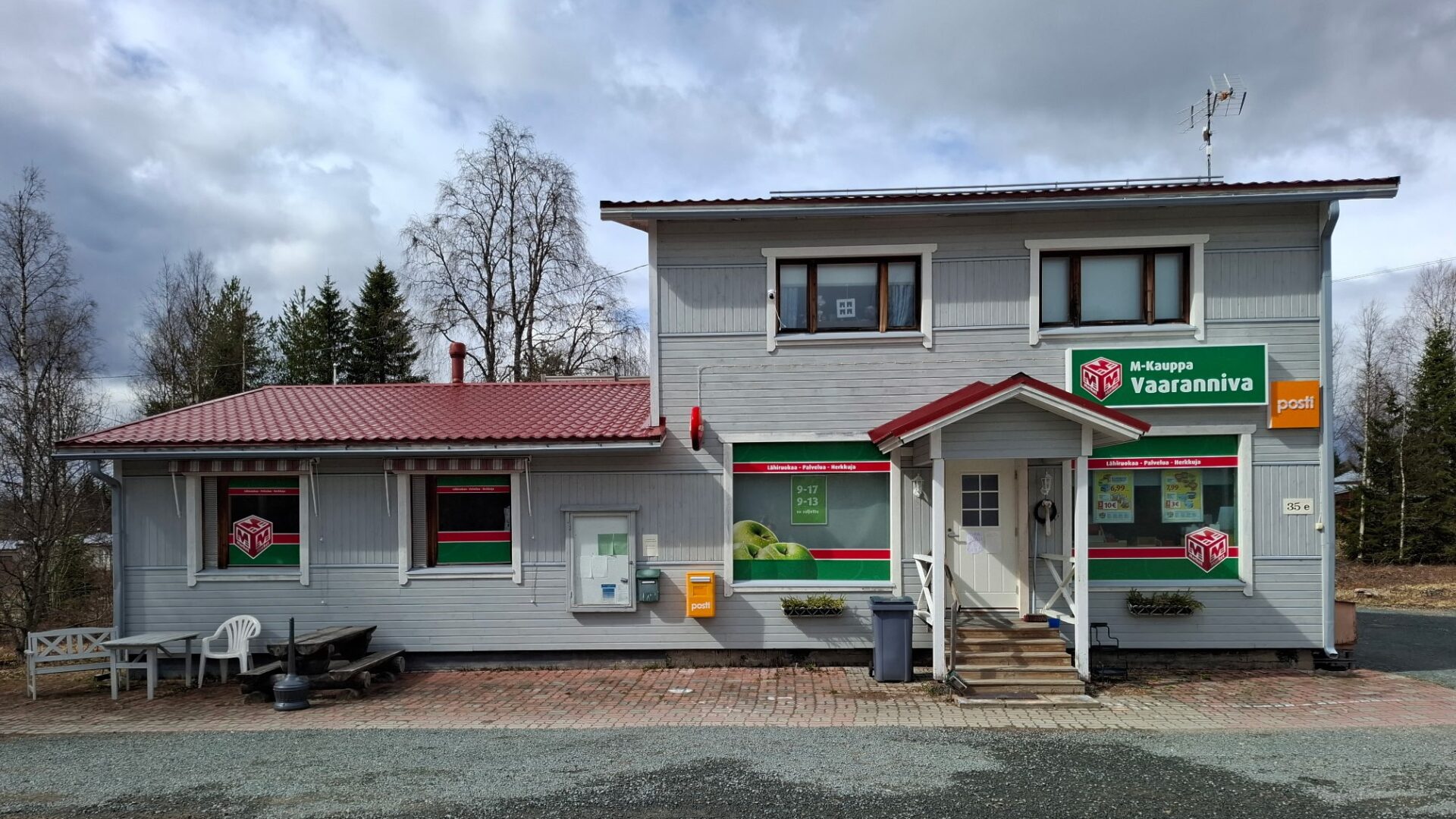 Vaaleanharmaa kyläkauppa, jonka seinällä postilaatikko ja pihalla pöytäryhmä