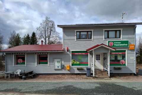 Vaaleanharmaa kyläkauppa, jonka seinällä postilaatikko ja pihalla pöytäryhmä