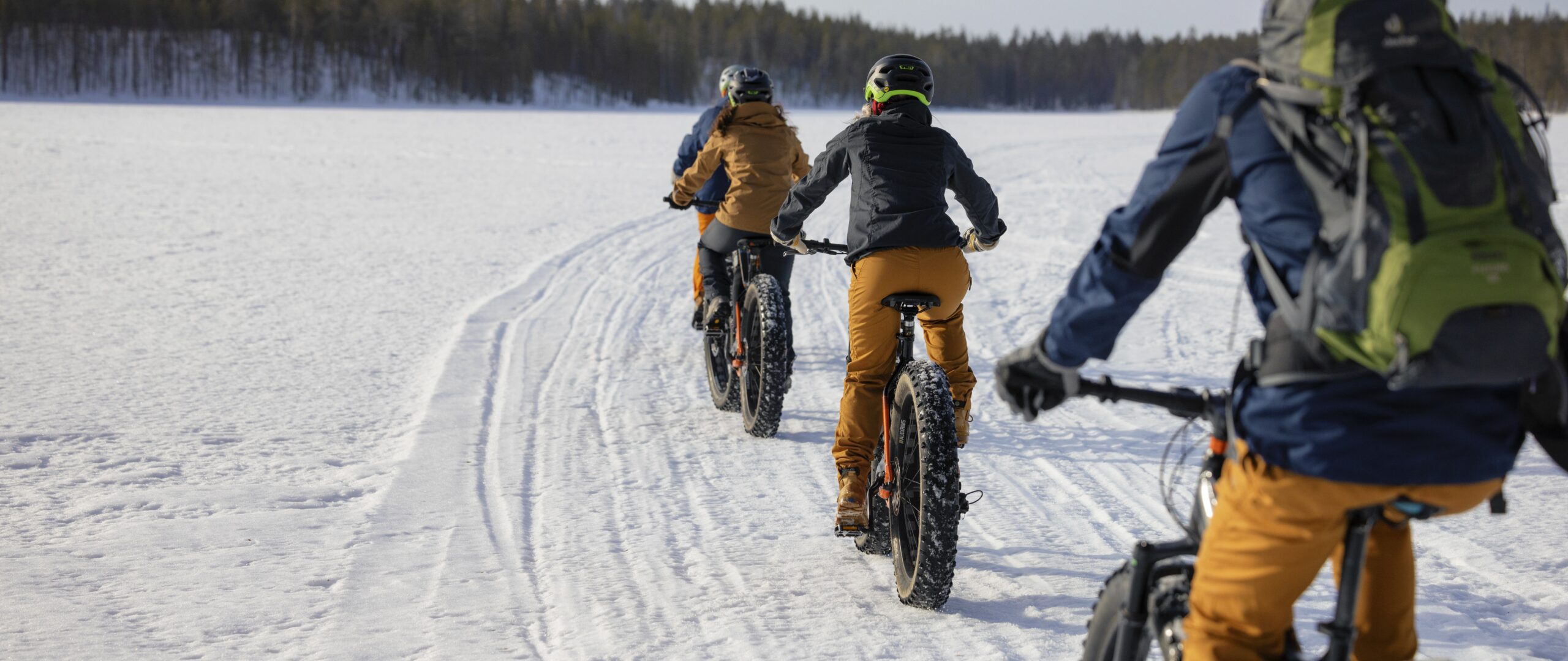 Joukko pyöräilee järvenjäällä auringoisessa talvimaisemassa.