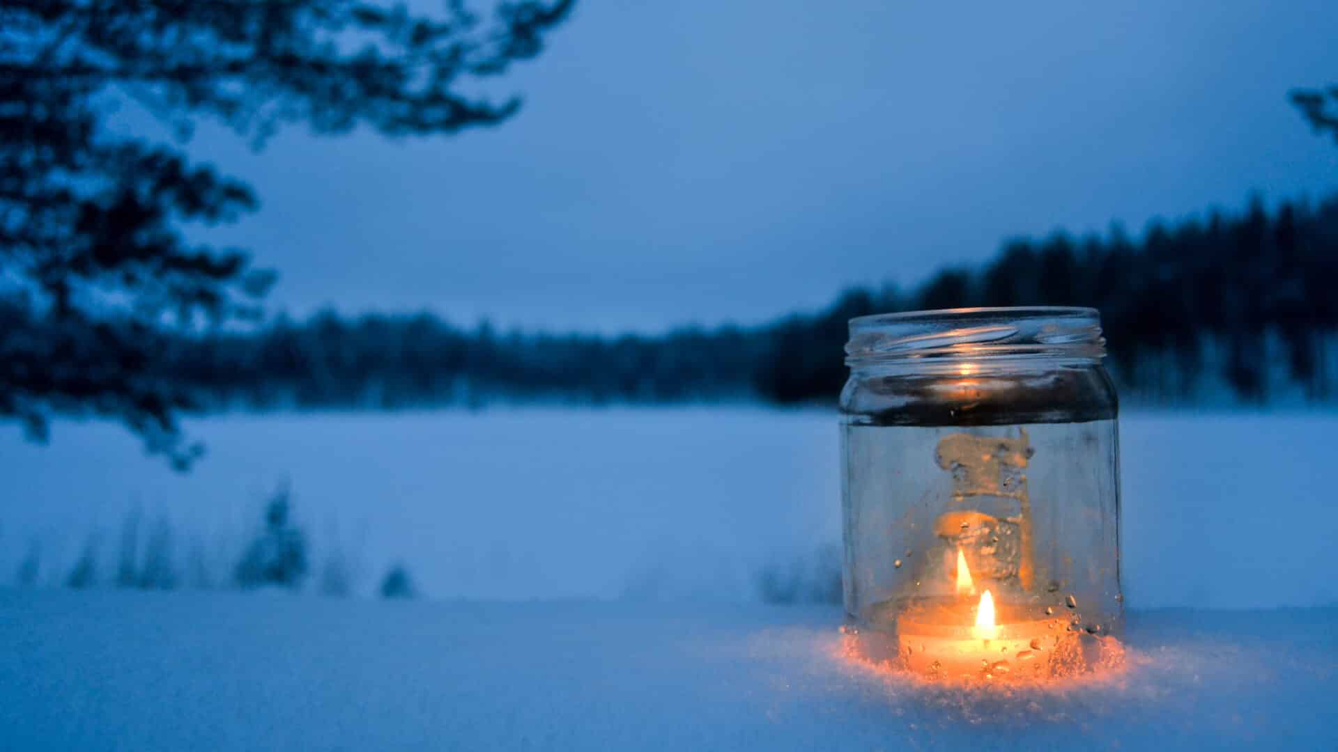 Kynttilä lasilyhdyssä lumisessa metsämaisemssa.