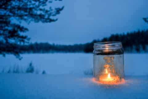 Kynttilä lasilyhdyssä lumisessa metsämaisemssa.