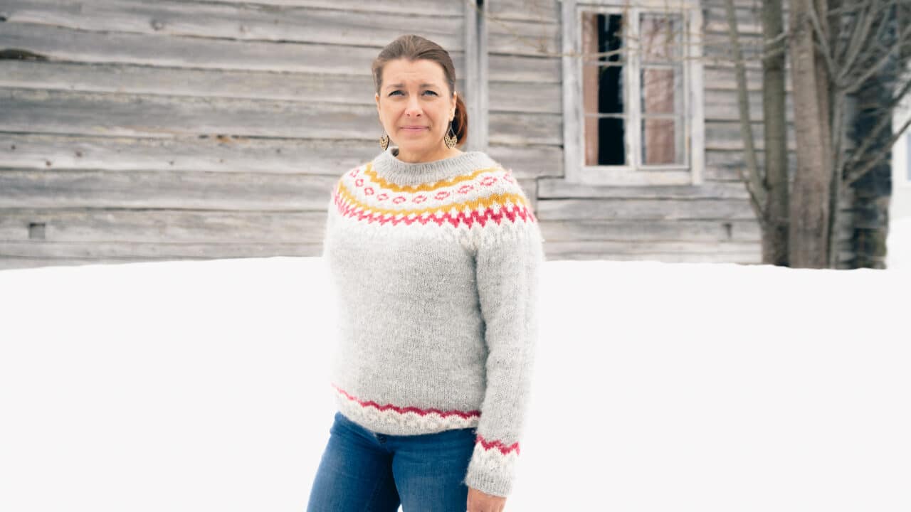 Ääninäyttelijä Mari Kela seisoo hirsirakennuksen edessä talvella, hänellä on päällään värikäs villapaita.