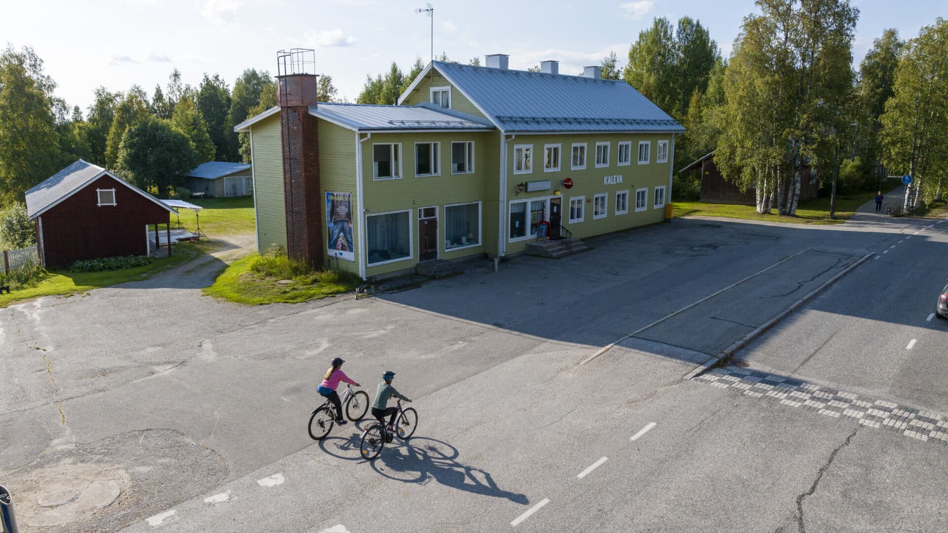 Pyöräilijät kesäiltana Suomussalmen kirkonkylällä. Kuva ilmasta kuvattu, Wanhan kalevan baari näkyy pyöräilijöiden takana. Kaleva on vihreä suuri rakennus.