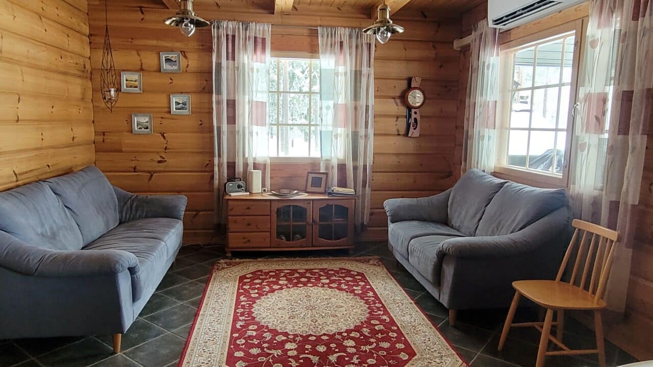 Olohuoneessa puna-valkoinen keskusmatto, siniharmaat sohvat, puinen kaappi.