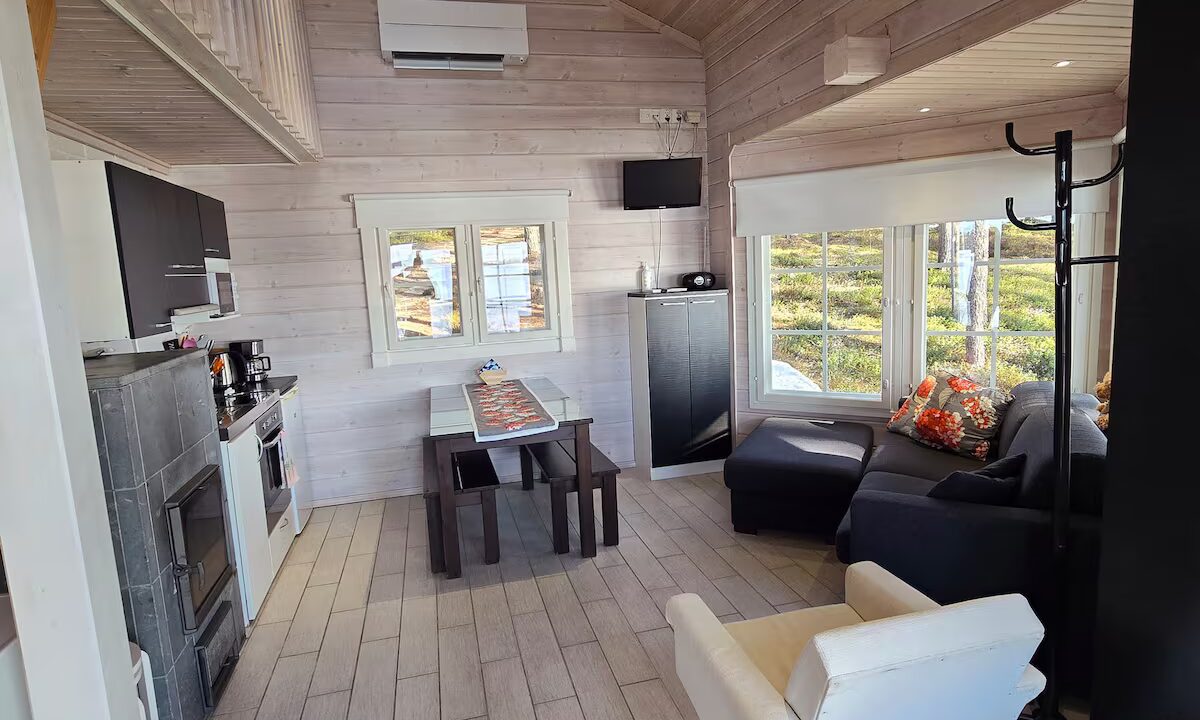 Vaaleassa, korkeassa tuvassa suuret ikkunat, mm. sohva, takka, nojatuoli ja ruokapöytä tuoleineen.