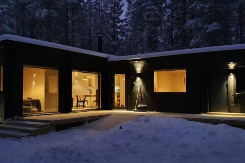 Musta mökki suurilla ikkunoilla talvimaisemassa.
