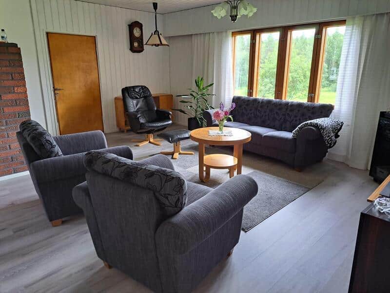 Olohuoneessa tummanharmaat sohva, nojatuolit ja matto sekä puinen pyöreä pöytä.