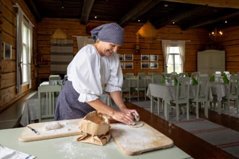 A cheerful woman is baking traditional Finnish flatbread (rieska). She is wearing a blue headscarf and smiling as she works the dough.